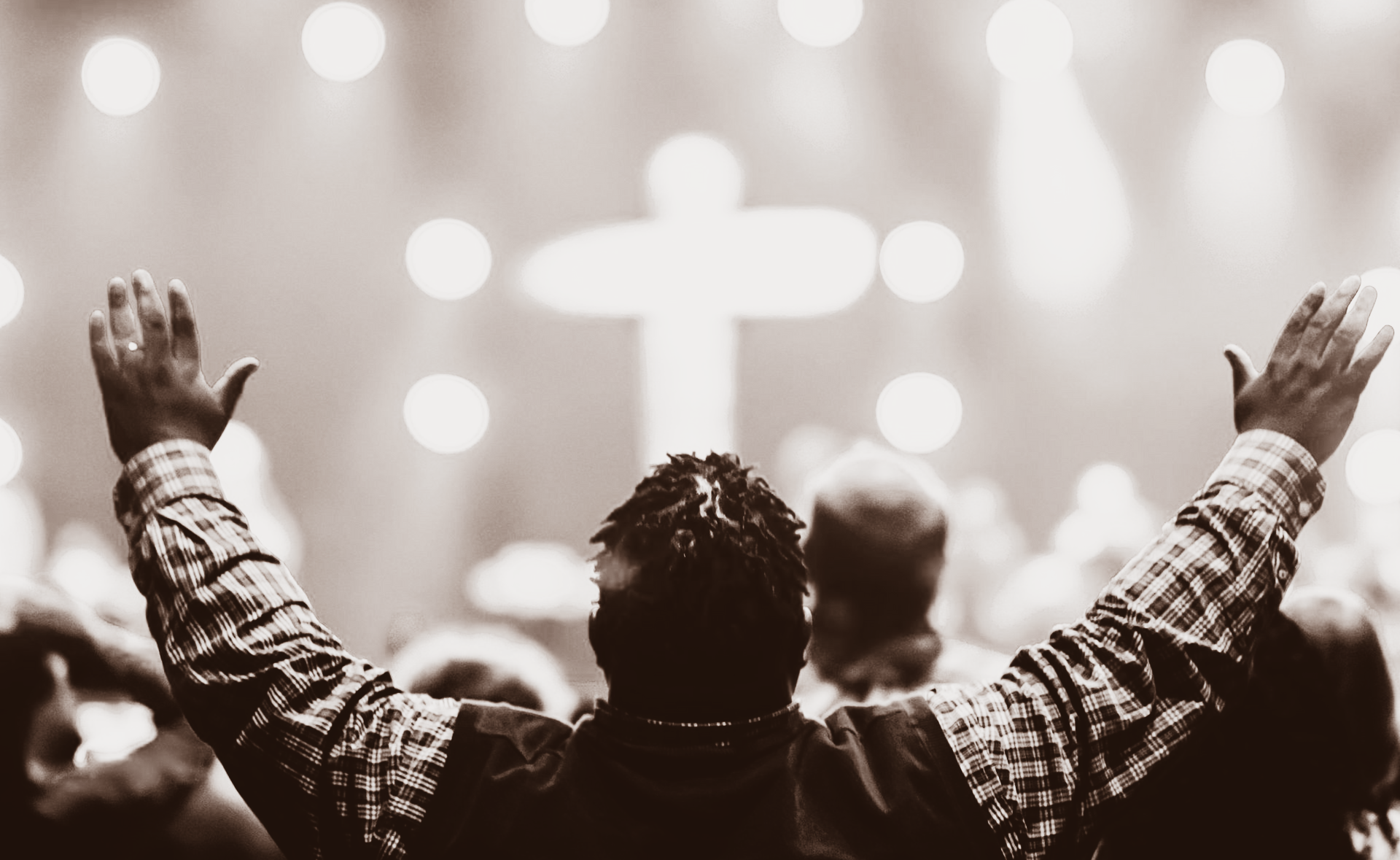 在拥挤的聚会中，一个人面对着明亮的十字形灯光，高举双臂进行礼拜。