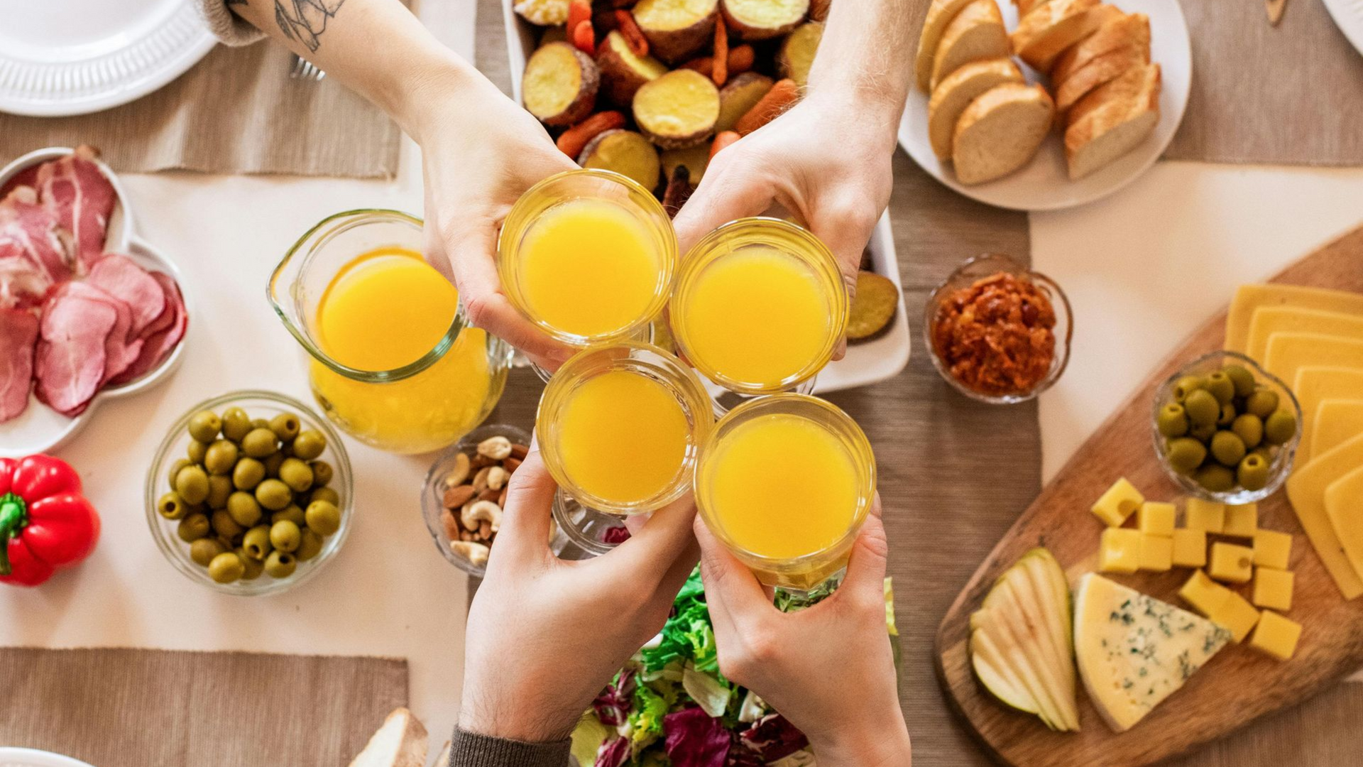 Four people toast with glasses of orange juice over a table filled with a variety of brunch foods.