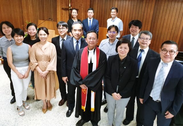 一群身着正装的人在一间镶有木板的房间里摆姿势拍照，其中一人身穿神职人员的法衣，站在房间中央。