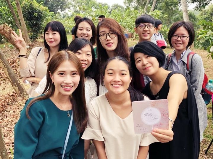 一群人在户外摆姿势拍照，其中一人拿着一张小卡片，所有人都面带微笑，背景是树林。