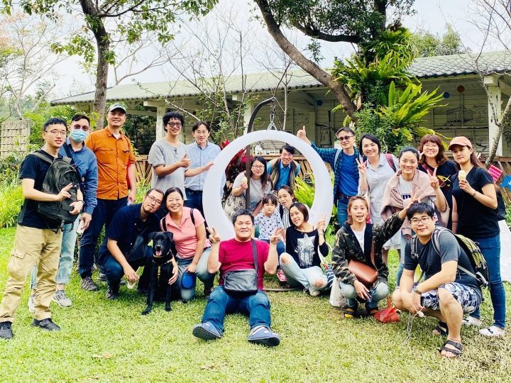 一群人在绿草茵茵、宛如公园般的环境中，围绕着一个巨大的白色圆形相框摆姿势拍照。