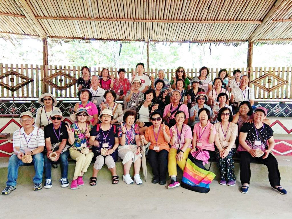 一大群人坐在茅草屋顶的木结构建筑下，排成一排，面带微笑地拍摄集体照。