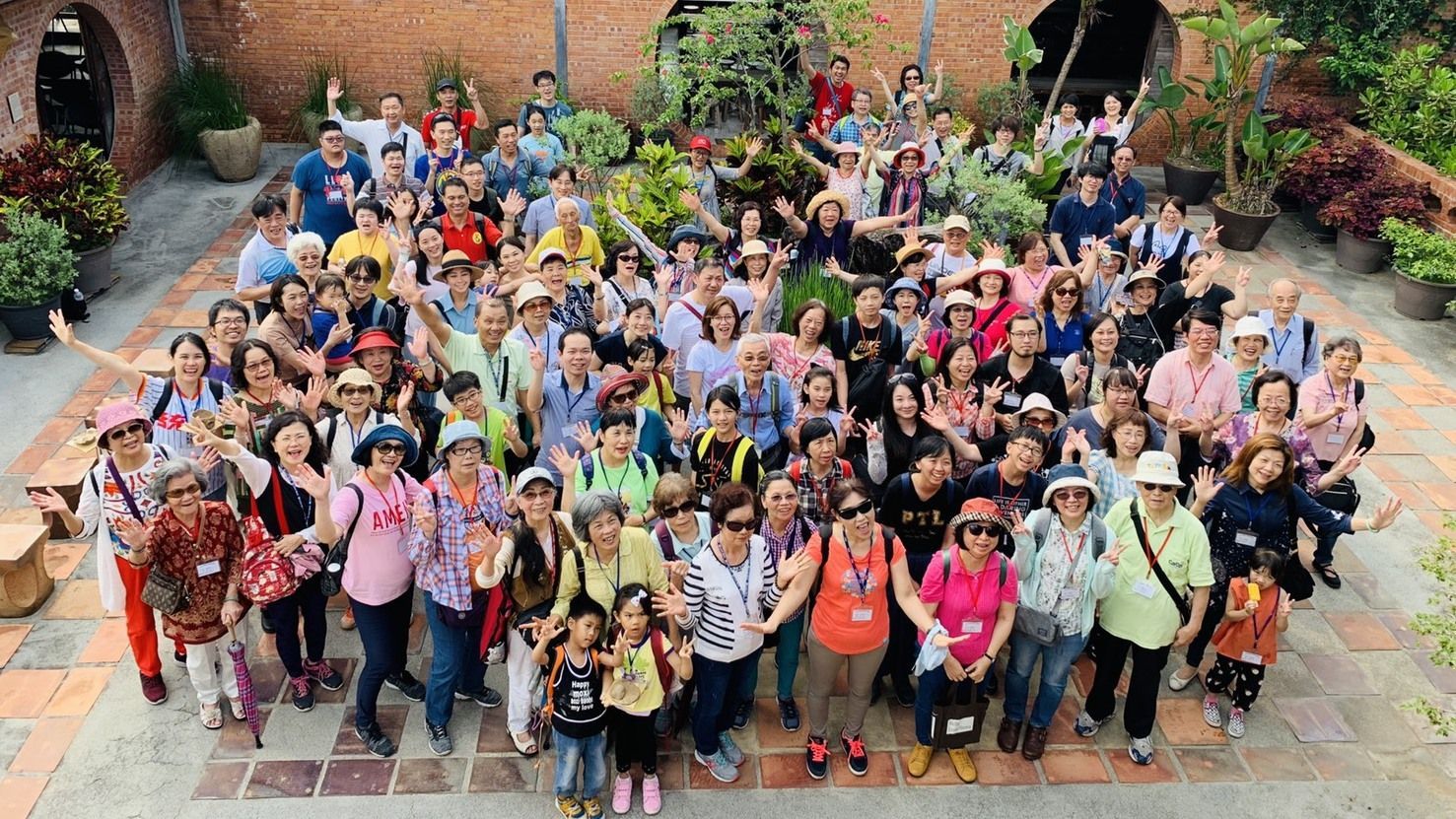 一大群不同背景的人们在砖墙环绕的户外庭院里摆姿势拍照，他们挥手微笑。