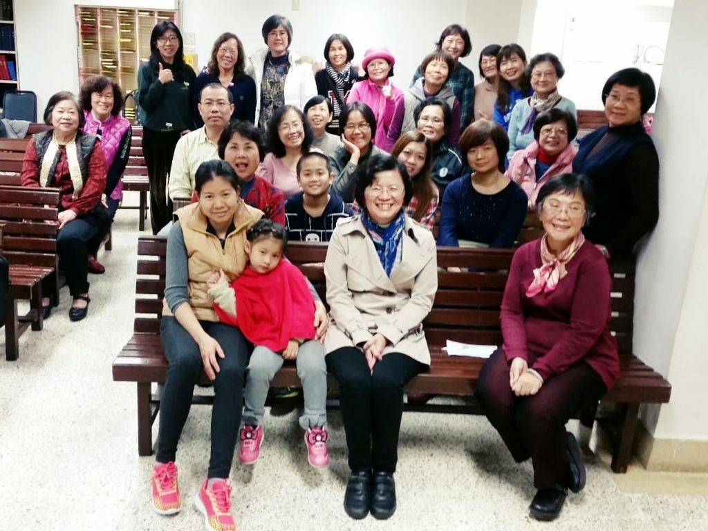 一大群人聚集在室内，许多人坐在木凳上，面带微笑地拍摄集体照。