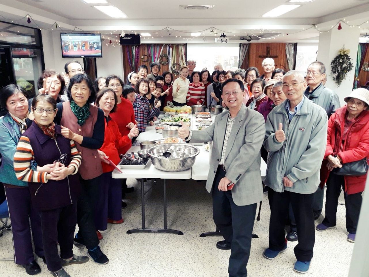 一群人站在社区活动中心的餐桌旁，桌上摆着几碗食物，对着镜头微笑。