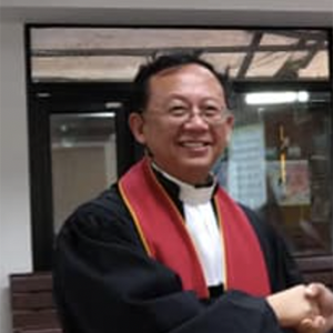 一位身穿黑色牧师长袍、披着红色圣带的人面带微笑，在室内与人握手。