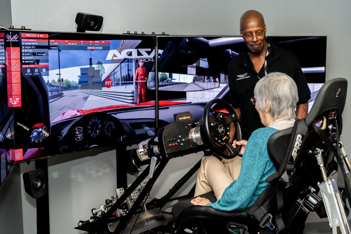 An elderly woman is playing a video game in a simulator while a man watches.
