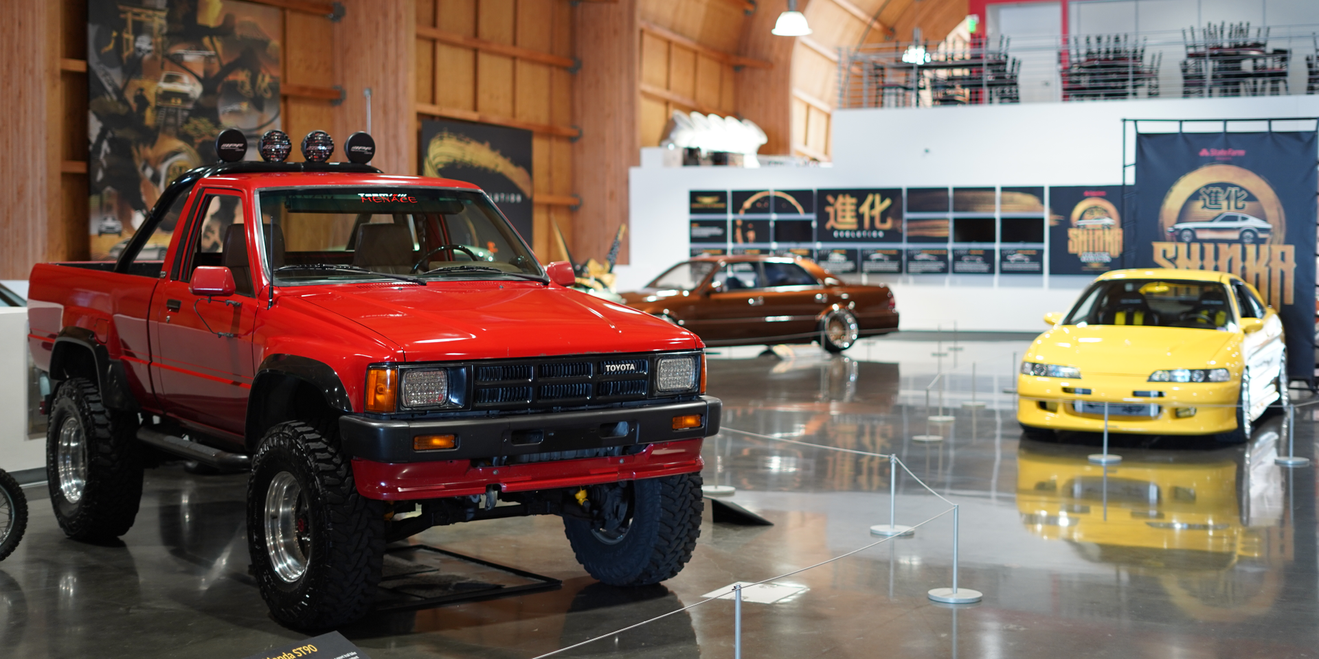 A red truck is parked in a garage next to a yellow car.