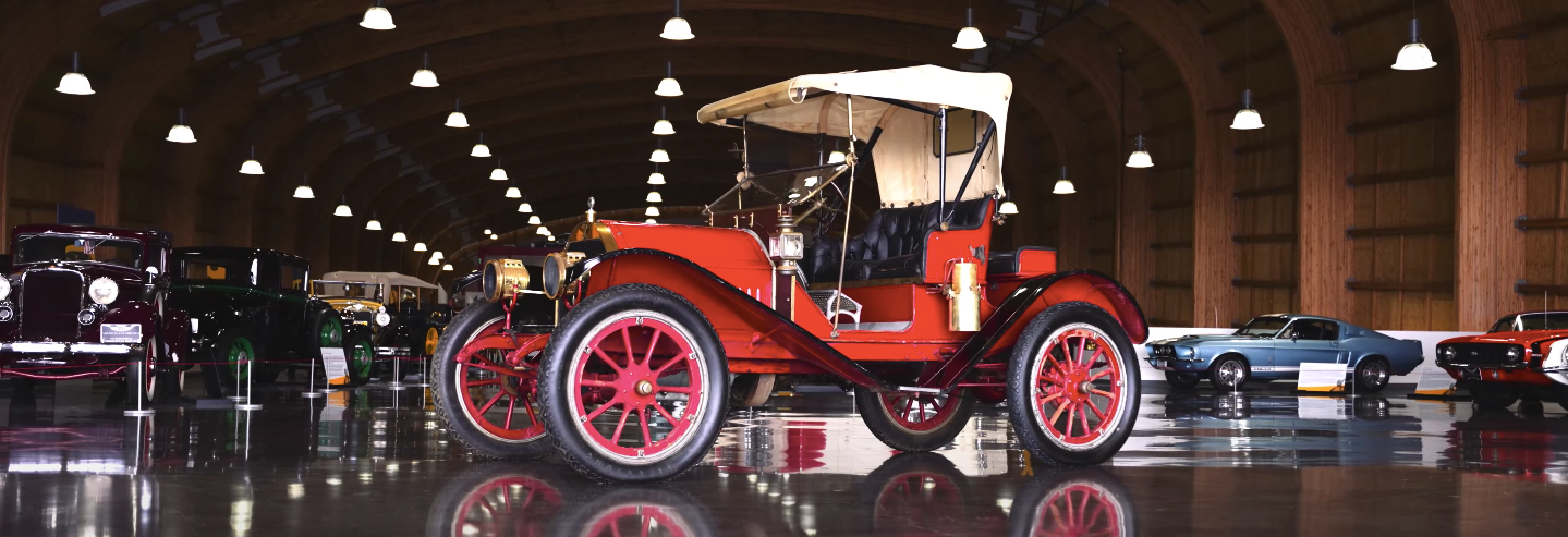 An old red car is parked in a garage with other cars.