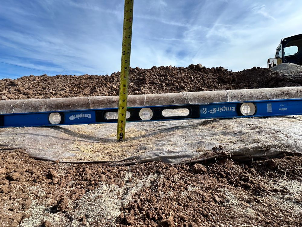 A level is used to measure ground covered with landscape fabric; dirt and sky in background.
