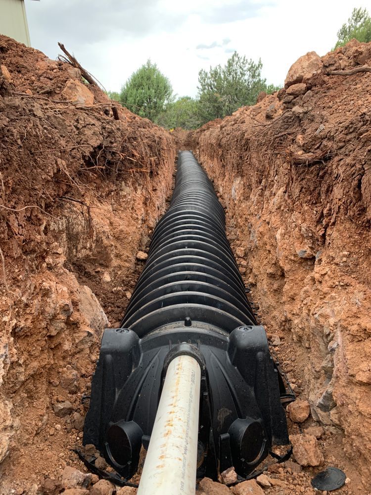 A black corrugated drain pipe installed in a trench filled with reddish-brown soil, ready for burial.
