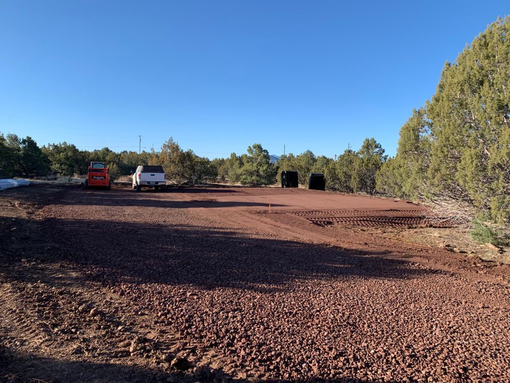 A large cleared area covered in red gravel, with vehicles and trees under a blue sky.