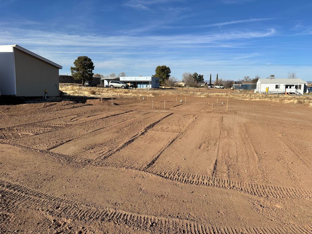 Dirt field with tractor tracks, a few buildings, and blue sky.