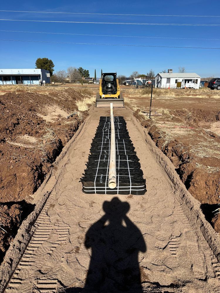 Construction site: Mini excavator over a trench with an installed drain field, sandy soil, and buildings in the background.
