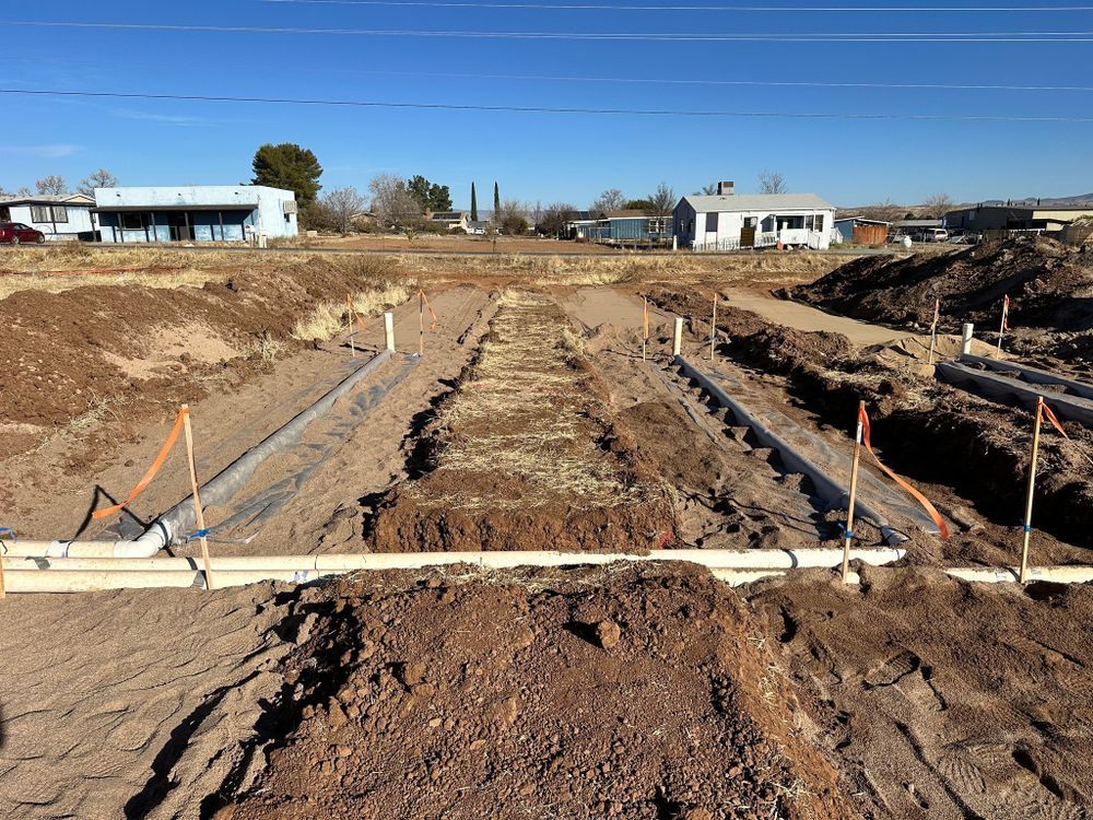 Excavated trenches with white pipes and orange markers, likely for underground utilities, in a dirt field. Buildings in the distance.