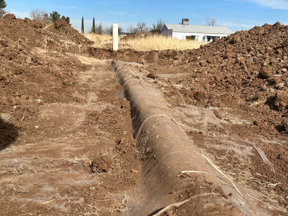 A trench with a brown pipe running through it, dug in reddish-brown soil, with a house in the background.