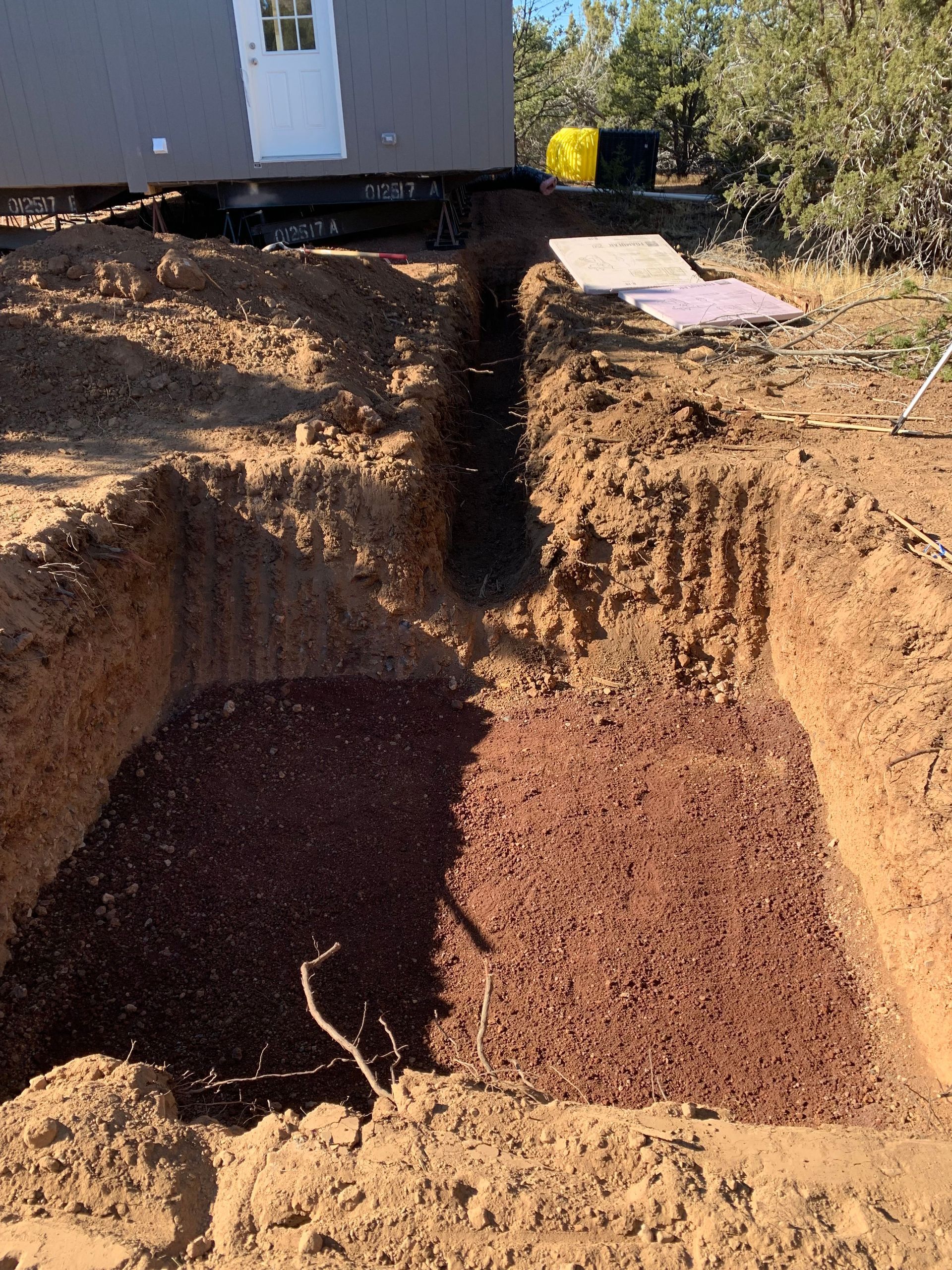 Excavation with red gravel base, ready for building foundation. Outdoors with a small structure in the background.