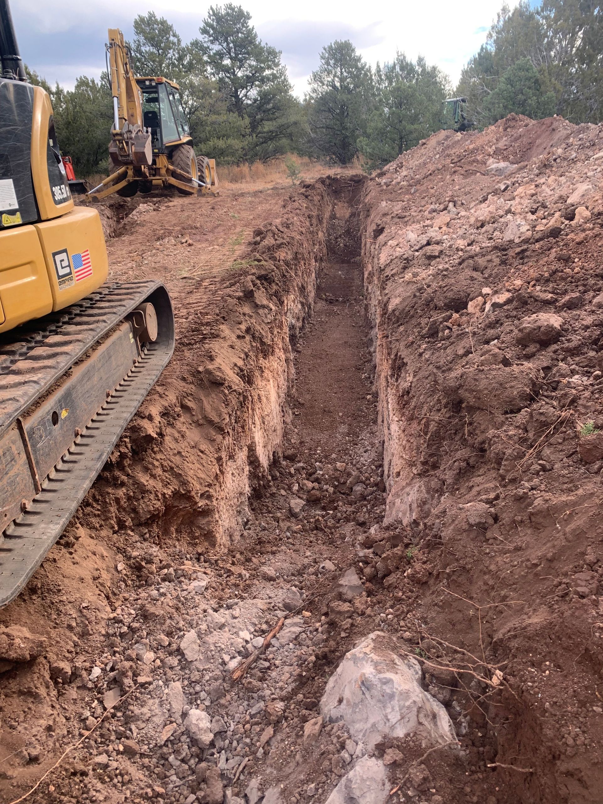 A long trench dug by a yellow excavator in a dirt field, trees in the background.