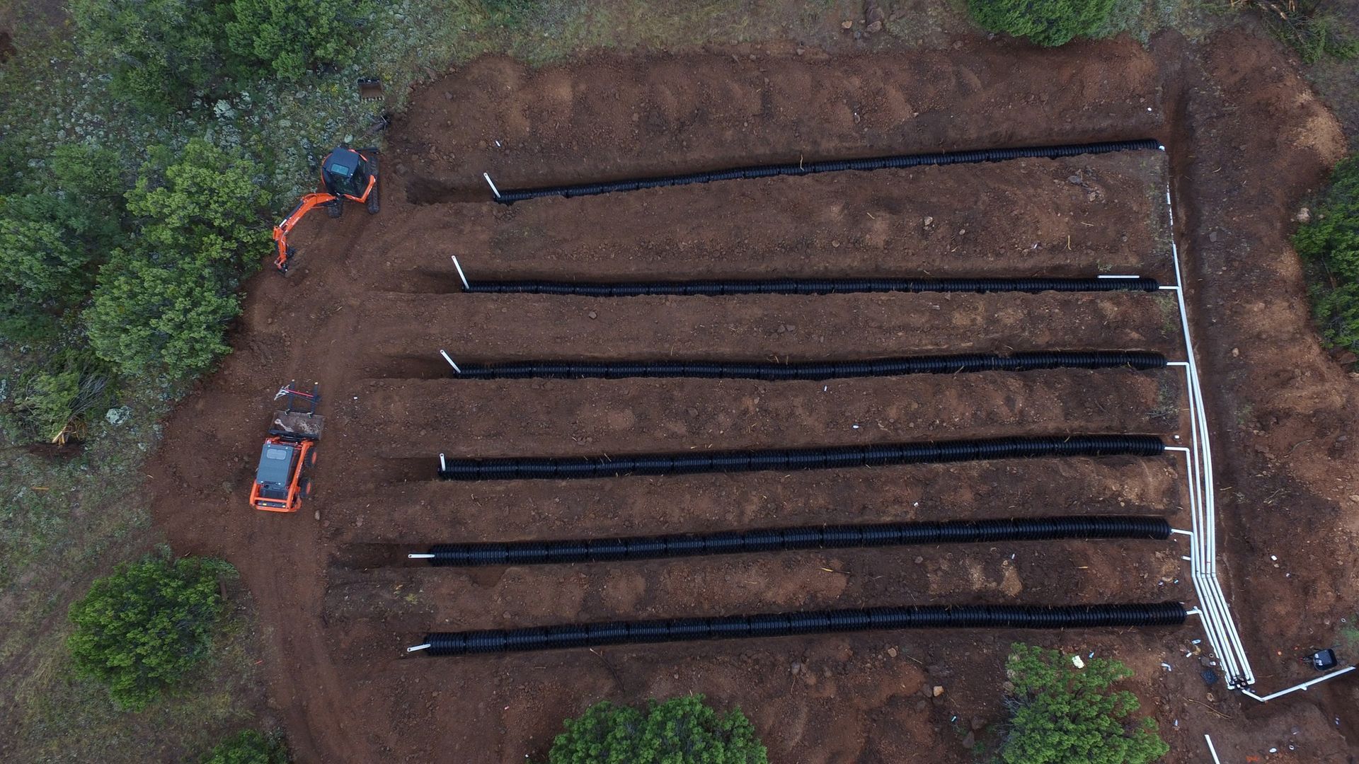 Aerial view: Construction site with parallel black pipes laid in trenches, two orange excavators.