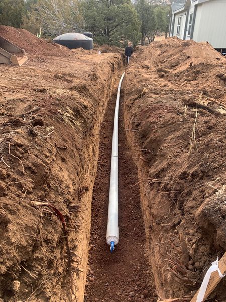 A narrow trench with white pipe laid in it; construction site. Brown dirt. A person in the background.