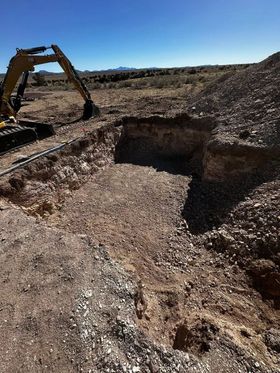 An excavator digs a large, open pit in arid, rocky terrain under a blue sky.