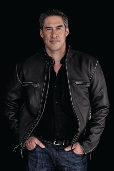 A professional headshot of author Brad Taylor. He is a man with short, salt-and-pepper hair and a friendly expression. He is wearing a blue patterned button-down shirt. The photo is taken outdoors with a soft, sun-drenched background of green foliage.