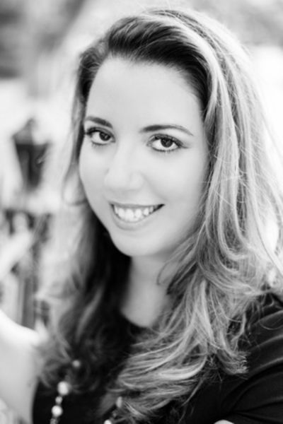 A professional black and white headshot of author Chanel Cleeton. She is a woman with long, dark wavy hair, smiling warmly at the camera. She is wearing a sleeveless dark top and a delicate gold necklace. The background is a soft-focus, bright outdoor setting with greenery and natural light.