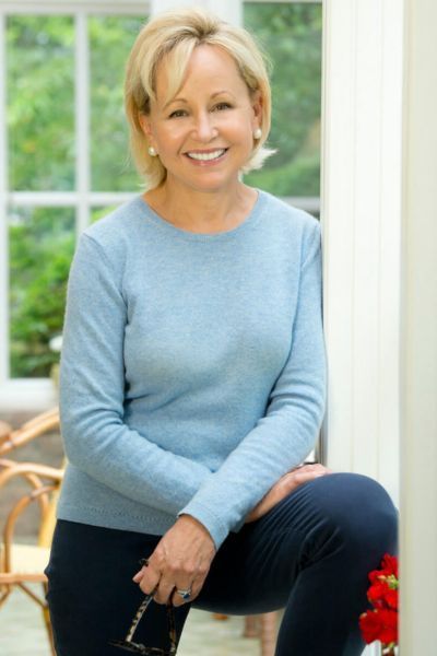A professional headshot of author Deborah Goodrich Royce. She is a woman with blonde, shoulder-length hair and a warm smile, wearing a dark navy blue blazer over a white top. She is posed against a soft-focus background of an indoor room with a bookshelf and warm lighting.