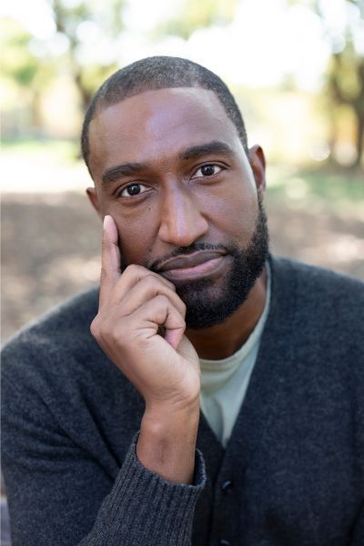 A professional headshot of author Jeff Boyd. He is a Black man with a short-trimmed beard and mustache, looking directly at the camera with a thoughtful expression. He has one hand resting against his cheek with his index finger pointed upward. He is wearing a dark charcoal gray cardigan over a light-colored shirt, set against a softly blurred outdoor background of trees and sunlight.