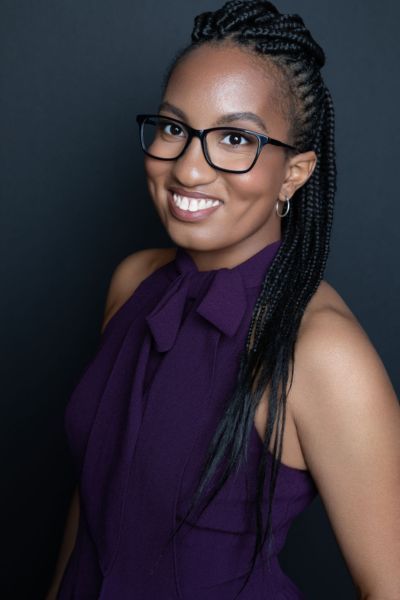 A professional headshot of author Nicole Glover. She is a Black woman with dark hair pulled back, wearing glasses and a dark blazer over a teal top. She is smiling warmly at the camera against a neutral, blurred indoor background.