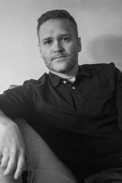 A professional black-and-white headshot of Oliver Munday. He is a man with short hair and a beard, wearing a dark button-down shirt. He is looking directly at the camera with a neutral, calm expression. The background is a simple, solid light color.