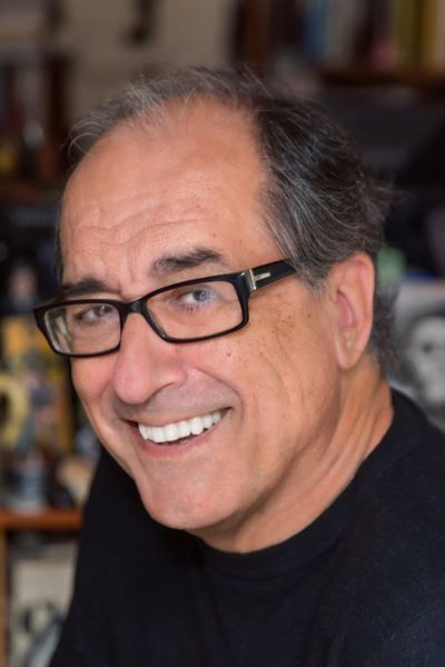 A professional headshot of author Scott Eyman. He is an older man with short, silver hair and glasses, smiling at the camera. He is wearing a dark suit jacket over a blue collared shirt. The background is a simple, out-of-focus indoor setting with warm lighting.