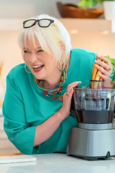 A candid, joyful photo of chef Lenore Nolan Ryan in a bright teal top. She is laughing while leaning over a kitchen counter, wearing her glasses perched on top of her blonde hair and colorful layered necklaces. She is playfully holding a bunch of whole carrots above the feed tube of a silver food processor.