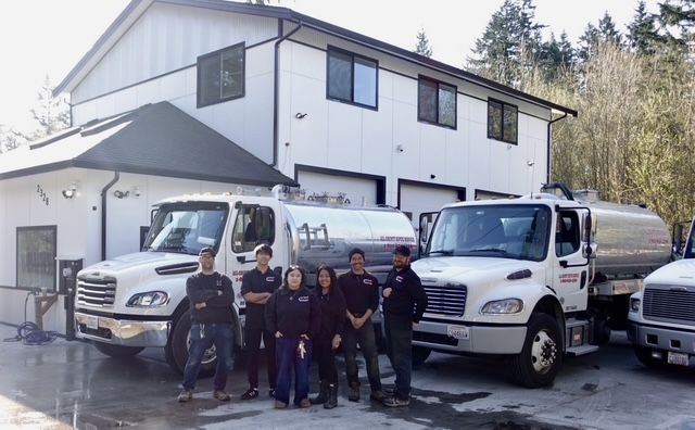 A group of six workers stand in front of two white tanker trucks parked outside a two-story industrial building.