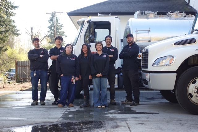 A group of seven people in dark uniforms stand in a row outdoors in front of a white truck and a large silver tank.