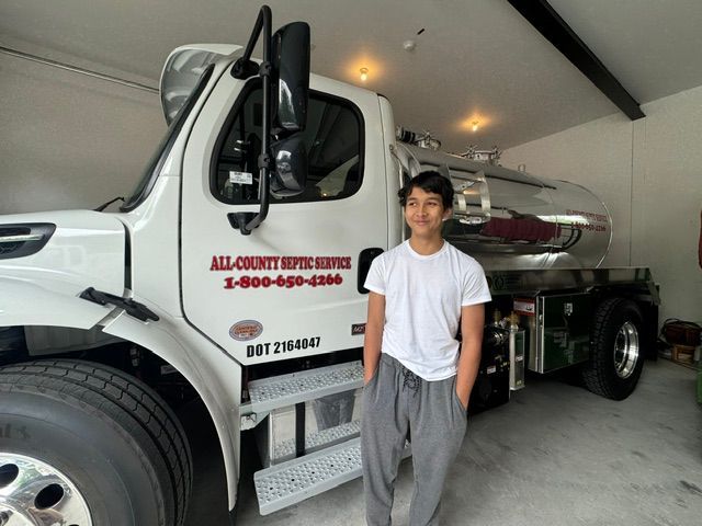 A young person stands in a garage beside a white septic service truck with company branding on the door.
