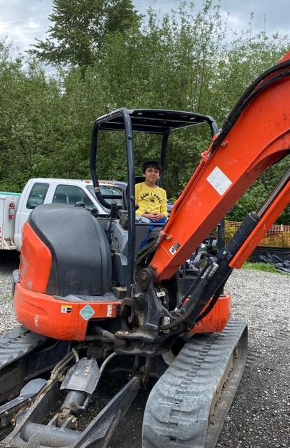 A person in a yellow shirt sits in the operator's seat of an orange mini-excavator parked on gravel.