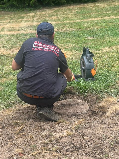 A technician in a black uniform kneeling in a grassy field to inspect a septic tank cover with specialized equipment.