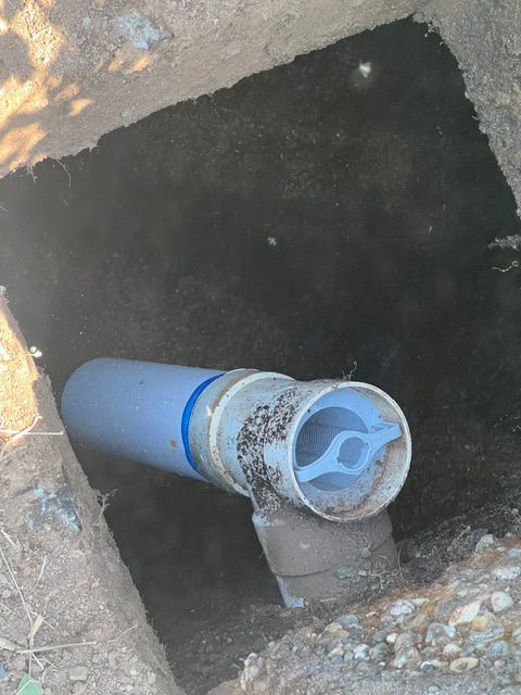 An open, dirt-filled ground hole revealing a blue and white plastic plumbing pipe with a butterfly-style handle valve.