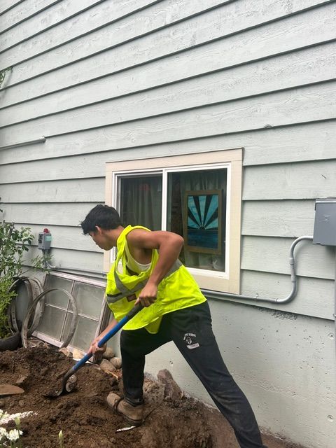 A person in a high-visibility yellow vest using a shovel to dig in dirt next to a house with light gray siding.