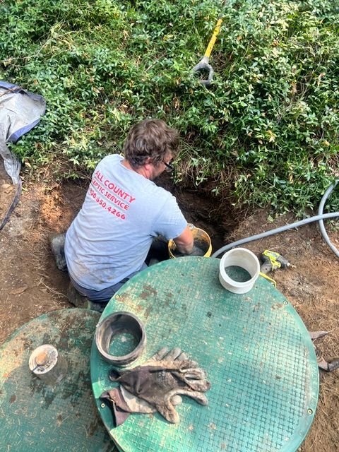 A service technician works on an exposed septic tank in a yard, with tools and a green access cover nearby.