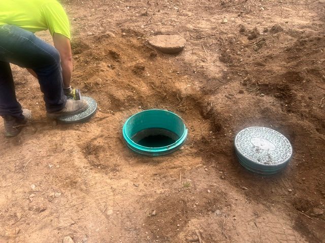 A person in a high-visibility shirt inspects an open, teal-rimmed septic tank riser next to a removed circular lid.