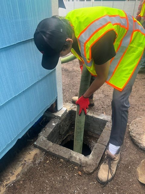 A worker in a high-visibility vest uses a green vacuum hose to clear an opening in the ground next to a blue building.