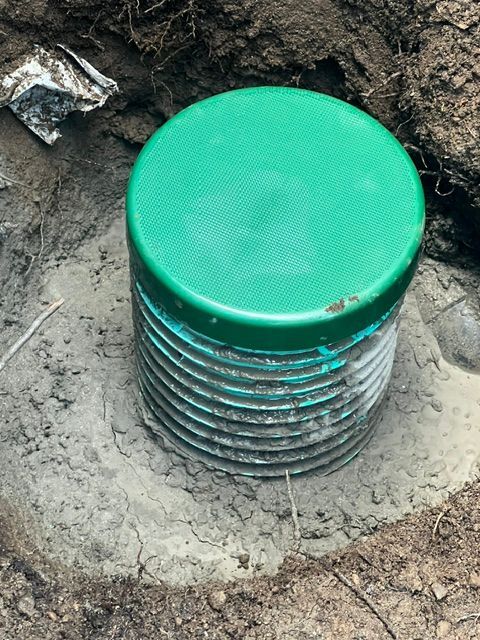 A green-capped, corrugated plastic drainage pipe installed in the ground and set in wet concrete.