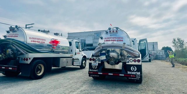 Two white tanker trucks parked on a gravel lot near industrial buildings, with a person in a safety vest visible nearby.