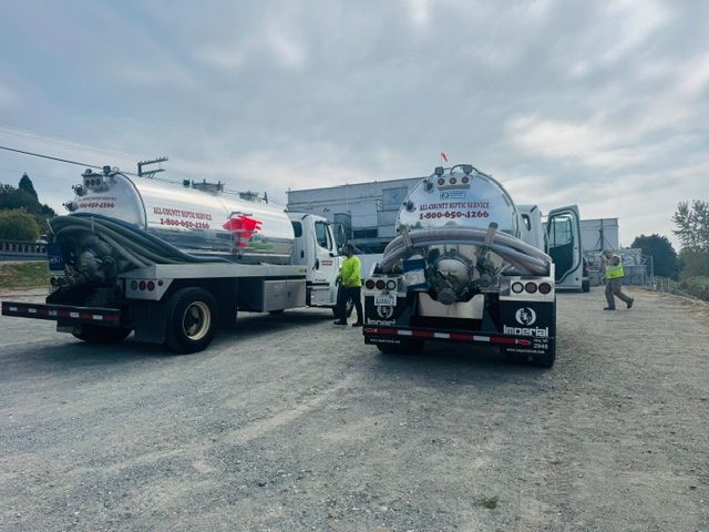 Two tanker trucks parked on a gravel lot with workers standing nearby under a cloudy sky.