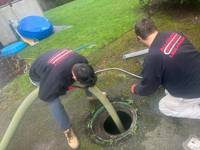 Two technicians in dark uniforms work together to pump out a septic tank via a hose inserted into an open ground access.