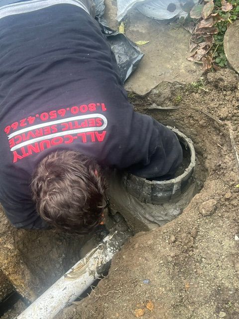 A service technician in a branded uniform leans into a hole in the ground to work on a septic system pipe.