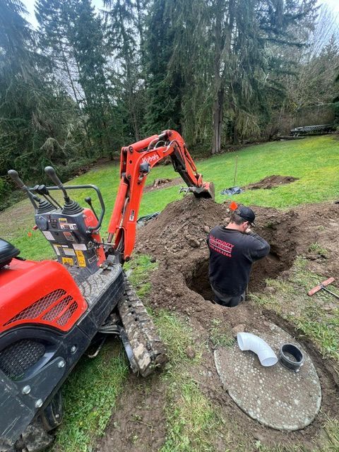 A worker stands in a trench next to an orange excavator in a grassy yard, with a pipe fitting nearby on the ground.