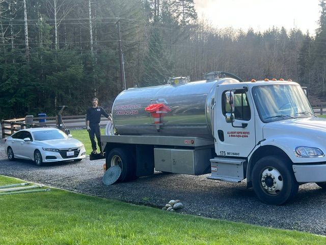 A person stands beside a large white tanker truck and a white sedan parked on a gravel driveway near a forested area.